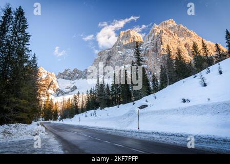 Leerer gerader Abschnitt einer Bergstraße am Fuße eines majestätischen schneebedeckten Felsgipfels an einem klaren Wintertag Stockfoto