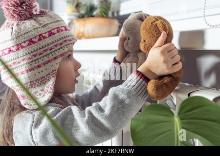 Nahaufnahme ruhiges, konzentriertes kleines Mädchen in gestricktem Hut und Pullover, das sich mit besten Freunden Stoffspielzeug in der Nähe des Heizkörpers aufwärmt, spielt und hat Stockfoto