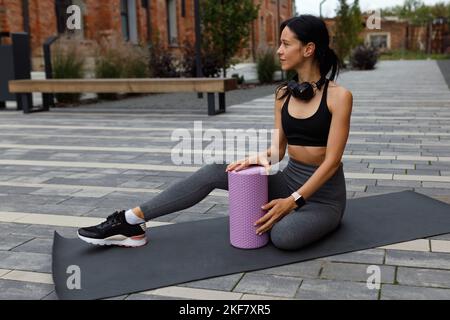 Die schöne weibliche Athletin mit Kopfhörern um den Hals sitzt auf einer Sportmatte mit einer Massagerolle Stockfoto