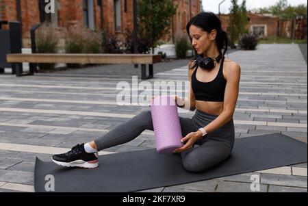 Die sportliche Frau mit einem Massageroller in den Händen sitzt auf einer Matte auf der Straße Stockfoto