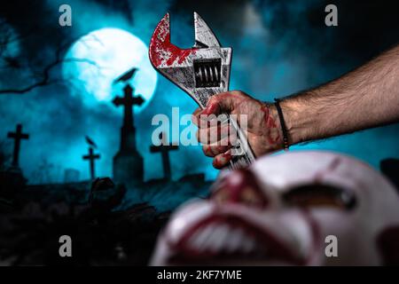 Eine blutige menschliche Hand hält ein Schraubenschlüsselblut mit einem Friedhof im Hintergrund und einer Clownsmaske vorne Stockfoto