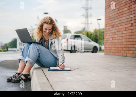 Internationaler Student, der online studiert, sich Notizen macht, während er einem Professor auf Videoanruf folgt und im Freien sitzt. Konzept von Bildung, Campus, Teenager Stockfoto