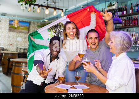 Fröhliche, vielfältige Fußballfans, die die Flagge Mexikos halten und in der Sportbar Bier genießen Stockfoto