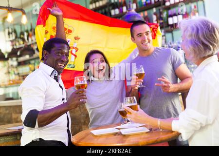 Fröhliche internationale Fußballfans winken mit spanischer Flagge und trinken Bier in der Sportbar Stockfoto