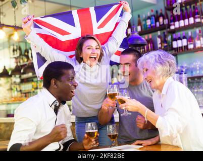 Fröhliche Sportfans, die die Flagge Großbritanniens halten, den Sieg der Nationalmannschaft feiern und im Pub alkoholische Getränke trinken Stockfoto