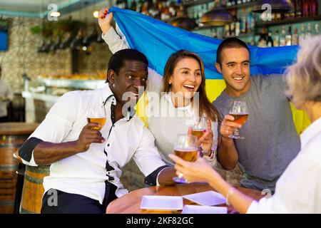 Glückliche internationale Sportfans winken mit ukrainischer Flagge, während sie Fußballspiele gucken und im Pub Bier trinken Stockfoto