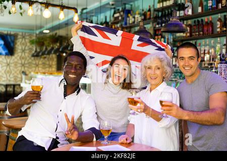 Glückliche multirassische Freunde, die die Flagge Englands halten, sich über den Gewinn eines Fußballspiels freuen und in der Bar Bier trinken Stockfoto