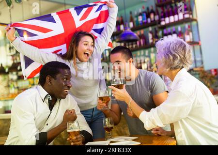 Englische diverse Fußballfans mit Flagge feiern den Sieg der Nationalmannschaft mit einem Pint Bier im Pub Stockfoto