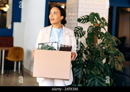 Eine begeisterte, erfolgreiche, motivierte brasilianische oder hispanische Frau kommt in ein neues Arbeitsbüro, hält einen Karton mit Arbeitsattributen in den Händen, inspiziert den Arbeitsplatz erstaunt und lächelt glücklich Stockfoto