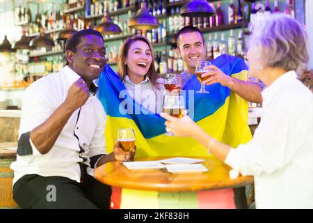 Glückliche internationale Sportfans winken mit ukrainischer Flagge, während sie Fußballspiele gucken und im Pub Bier trinken Stockfoto