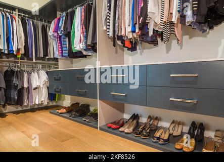 Begehbarer Kleiderschrank mit Schubladen, Herren- und Damenkleidung auf Kleiderbügeln, verschiedene Schuhe auf Regalen neben dem Hauptschlafzimmer in einem luxuriösen, zeitgenössischen Haus. Stockfoto