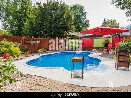 Unregelmäßig ovaler Swimmingpool im Boden, umgeben von braunen Pflastersteinen und umgeben von verschiedenen tropischen Pflanzen und Blumen im Garten. Stockfoto