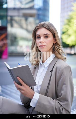 Eine junge Frau im Firmenanzug sitzt mit Notizbuch und Stift, notiert sich, arbeitet und schreibt ihre Ideen auf Stockfoto