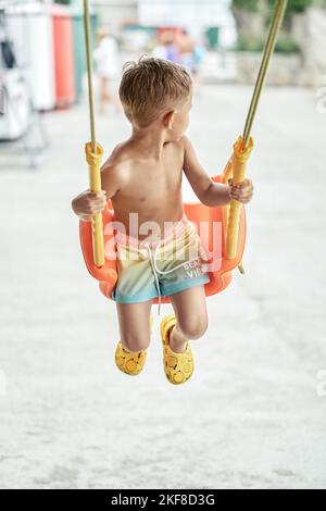 Kleiner blonder Junge schwingt auf einer orangen Schaukel und blickt zurück. Kleine Kinder schaukeln auf dem Kinderspielplatz des Hotels und genießen einen schönen Urlaub am Meer Stockfoto