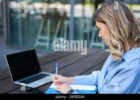 Nahaufnahme eines Porträts einer Studentin, die an Online-Kursen teilnimmt, mit einem Laptop an der frischen Luft sitzt und sich Notizen macht. Videochat für Geschäftsfrau Stockfoto