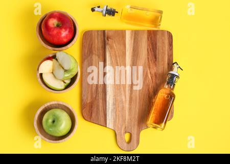 Zusammensetzung mit Flaschen Apfelessig, Früchten und Holzbrett auf gelbem Hintergrund Stockfoto