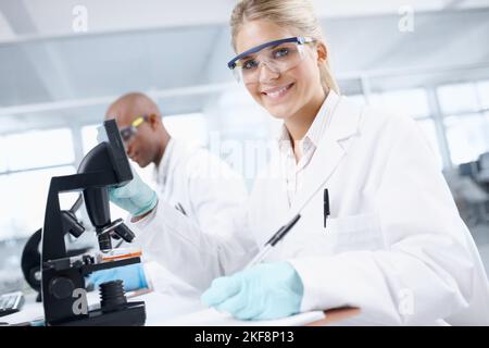 Forschen, um Wissen zu gewinnen. Porträt einer wunderschönen Wissenschaftlerin vor einem Mikroskop mit einem männlichen Kollegen im Hintergrund. Stockfoto