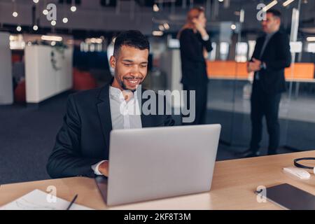 Lächelnder afrikanischer Geschäftsmann, der in einem modernen Büro mit Hintergrund von Kollegen einen Laptop arbeitet Stockfoto