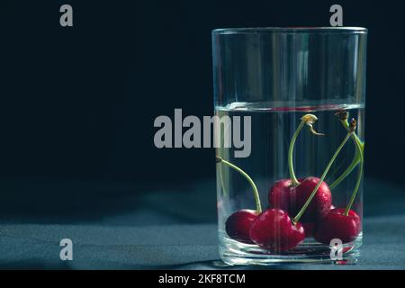 Kirschen in einem Glas Wasser auf schwarzem Hintergrund auf dem Tisch, Obst Hintergrund, Früchte Stockfoto