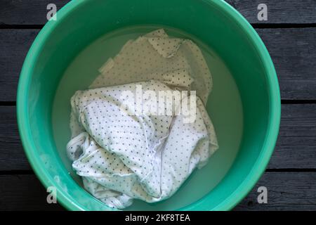 Weißes T-Shirt, waschbar in Wasser in einer grünen Kunststoffschüssel auf einem schwarzen Holzboden, Handwäsche in einer Schüssel Stockfoto