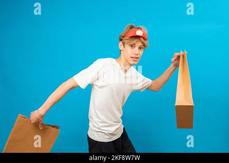 Harry auf. Junge Verkäuferin im lässigen weißen T-Shirt mit Papiertasche läuft auf blauem Hintergrund Studio-Porträt. Menschen Lifestyle-Konzept. Teen hält Packa Stockfoto