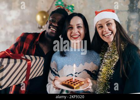 Frau hält Geburtstagskuchen Vorbereitung Kerze blasen, Freunde um küssen Wangen Stockfoto