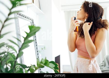 Hübsche Frau aus gemischter Rasse, die ihr lockiges Haar am Fenster zu Hause mit Feuchtigkeitsspray besprüht. Afro america weiblich, die Ölspray für ihre Herrin verwendet. S Stockfoto