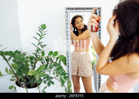 Hübsche Frau aus gemischter Rasse, die ihr lockiges Haar am Fenster zu Hause mit Feuchtigkeitsspray besprüht. Afro america weiblich, die Ölspray für ihre Herrin verwendet. S Stockfoto