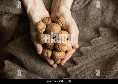 Eine alte Frau, die zu Hause viele Walnüsse in den Händen hält, auf dem Hintergrund eines braunen Schals, Nüsse in den Händen der Großmutter, gesundes Essen, veganes Essen Stockfoto