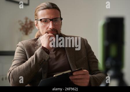 Ernsthafter Psychotherapeut in Brillen, der dem Patienten zuhört, während er mit Clipboard im Sessel im Büro sitzt Stockfoto