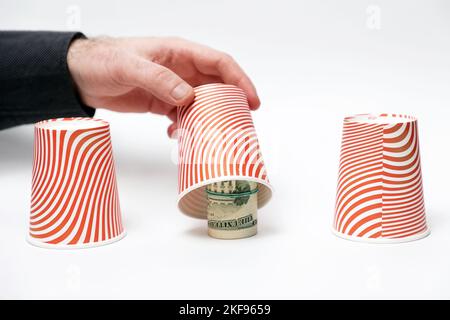 Geldbetrug. Nahaufnahme der männlichen Hand öffnet den Papierbecher und zeigt zusammengerollte Dollar. Weißer Hintergrund. Konzept von Betrug, Glücksspiel und Aktienmarkt. Stockfoto