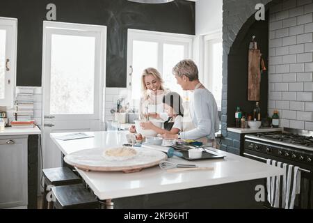 Großmutter, Mutter und Kind backen in der Küche zusammen, während sie sich im Familienhaus zusammenbinden. Ältere Frau, Mutter und Mädchen, die Essen zum Abendessen kochen Stockfoto