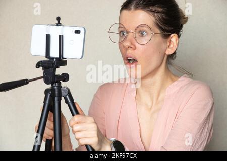 Das Mädchen schaut überrascht auf das Telefon auf einem Stativ zu Hause am Tisch, überrascht und erfreut auf dem Gesicht des Mädchens Stockfoto