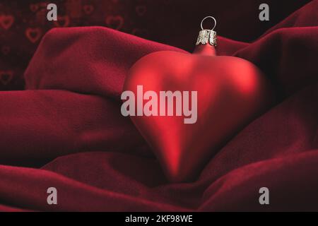 Eine rote weihnachtskugel in Herzform liegt auf einer weichen Samtdecke und glänzenden kleinen Herzen im Hintergrund. Stockfoto