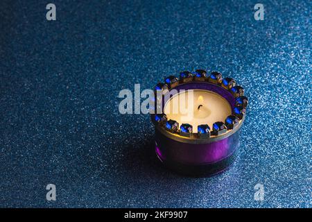 Eine brennende Teelicht Kerze in einem lila Glashalter auf einem dunkelblauen Hintergrund mit Glitzer. Stockfoto