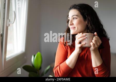 Junge glückliche, schöne Frau, die eine Tasse heißes Getränk in der Hand hält und durch das Fenster schaut und sich wohlfühlt Stockfoto