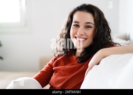Junge schöne Brünette Frau lächelt vor der Kamera, während sie sich auf der Couch zu Hause ausruhen Stockfoto