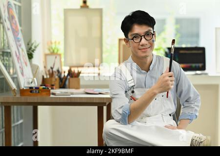 Porträt eines hübschen männlichen Künstlers, der den Pinsel in der Hand hält und vor der Kamera lächelt, während er in einem hellen Kunststudio sitzt Stockfoto