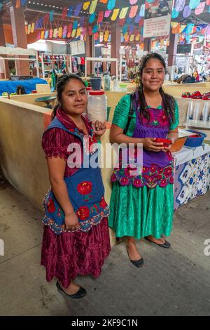 Zwei junge Zapoteerinnen aus San Marcos Tlapazola auf dem Markt in Tlacolula, Oaxaca, Mexiko. Sie tragen traditionelle Satinkleider und sticken Stockfoto