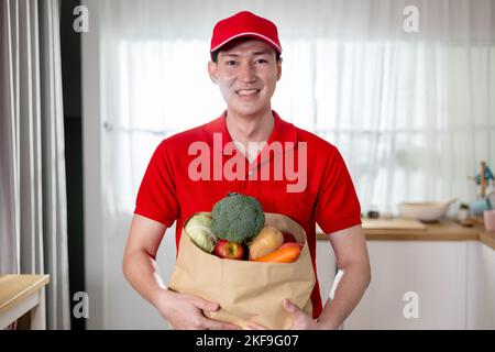 Lächelnder asiatischer Lieferer in roter Uniform, der Lebensmittel in einer Papiertüte trägt und Lebensmittel, Obst und Gemüse zu Hause liefert Stockfoto
