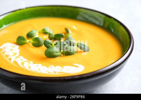 Nahaufnahme der Butternut-Kürbiscremesuppe, garniert mit schwerer Creme und frischer Sonnenblumen-Microgreen. Cremige Kürbis-Fallsuppe in grüner Schüssel dagegen Stockfoto