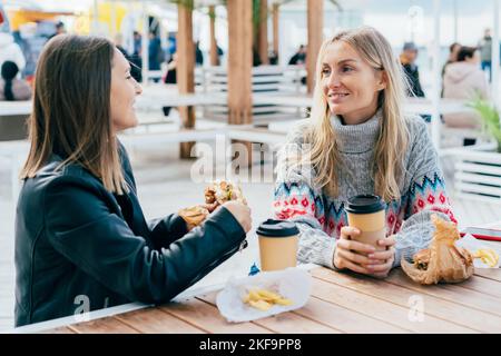 Zwei Freundinnen essen Hamburger, trinken Kaffee und plaudern auf der Straße, sitzen am Tisch. Stockfoto