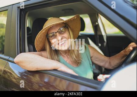 Fröhliche ältere Fahrerin im Strohhut, die in einem neuen Auto sitzt und lächelt und die Kamera anschaut, die die Reise genießt. Fahrkurse und Lebensversicherung. Aktivitäten im Ruhestand und Roadtrip-Konzept Stockfoto