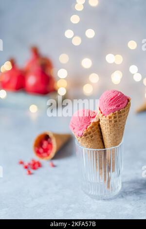 Gesundes, cremiges Granatapfel-Eis in einem Waffelkegel auf einem grauen Tisch, Hintergrund Girlanden Bokeh. Festliches veganes Dessert. Frohe Weihnachten. Stockfoto