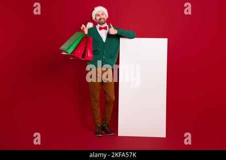 Ganzkörper-Porträt von eleganten Gentleman halten Pakete zeigen Daumen nach oben riesigen leeren Raum elektronische Panel auf rotem Hintergrund isoliert Stockfoto