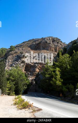Landstraße durch die typisch griechische mediterrane Landschaft mit Hügeln, Tannen und Büschen. Tourismus- und Urlaubskonzept. Rhodos-Insel, Griechenland. Stockfoto