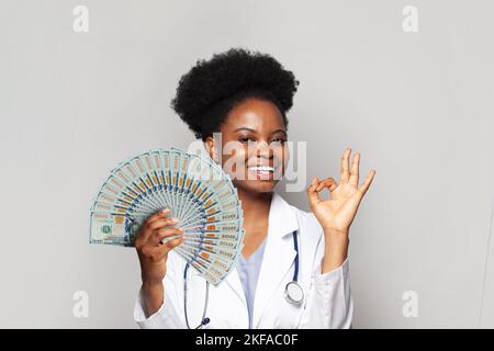 Erfolgreiche afroamerikanische Ärztin lächelt und zeigt OK und hält Geldfan vor weißem Hintergrund Stockfoto
