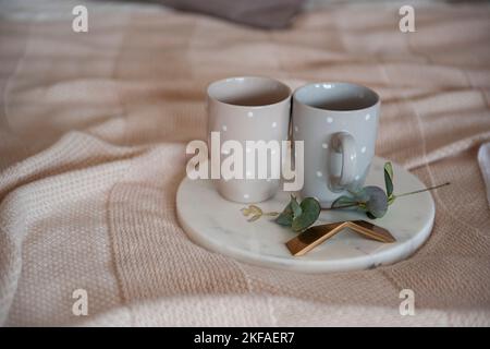 Auf einem gestrickten, strukturierten, weißen Karo liegt ein rundes Marmortablett mit einem Ast aus grünem Eukalyptus, auf dem sich zwei graue Tassen mit Tupfen befinden. Scannen Stockfoto