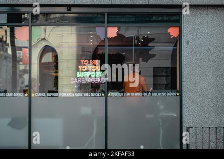 Die Mitarbeiter der Agentur arbeiten an einem Schaufenster mit Neonlicht und einem Mann, der am Telefon antwortet. Reflexionen im Fenster. Stockfoto