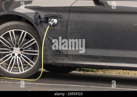 Schwarzes Hybridauto, das auf der Fahrbahn geparkt wurde und mit eingestecktem Strom aufgeladen wurde. Stockfoto